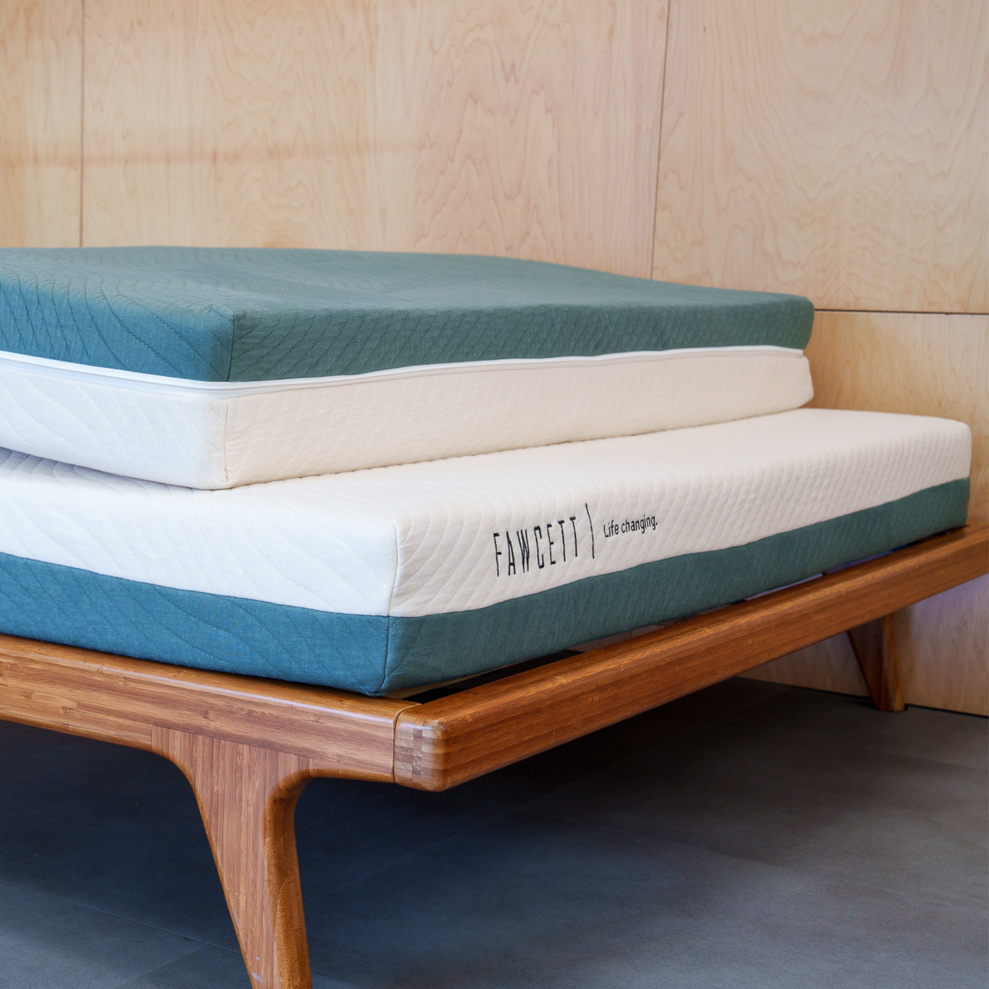 A folded Tofino mattress in teal and white on a wooden slat bed frame against a light wood panelled wall.