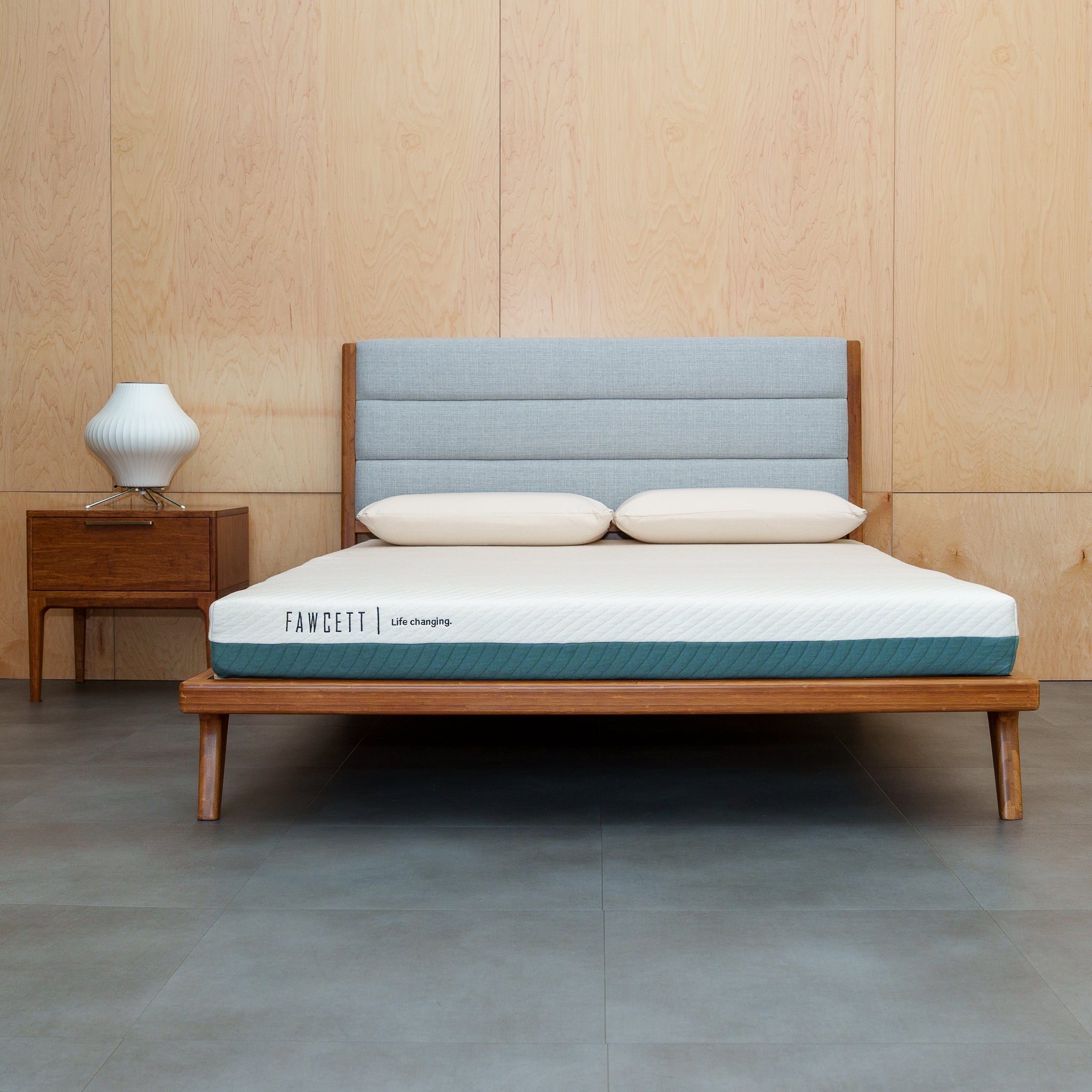 A tofino mattress on a wooden bed frame with a light gray upholstered headboard, styled in a bedroom setting with a wooden nightstand and lamp.