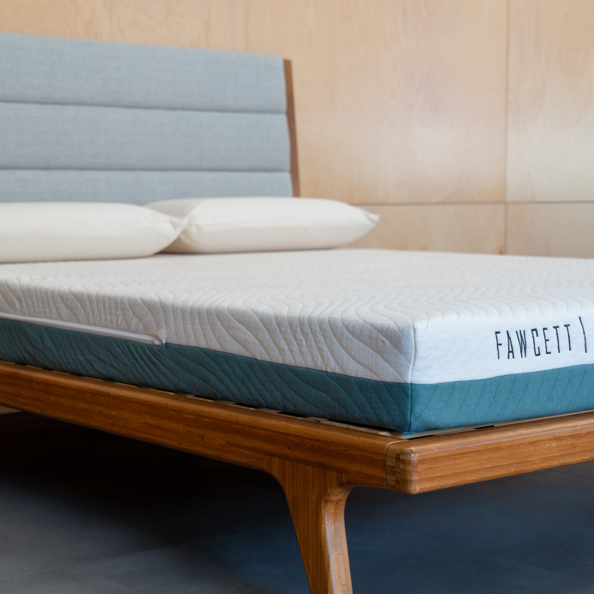 A close-up of a mattress on a wooden bed frame with a light blue upholstered headboard. The mattress has a white quilted top and a teal blue side panel, with the brand name 'FAWCETT' printed on the side.