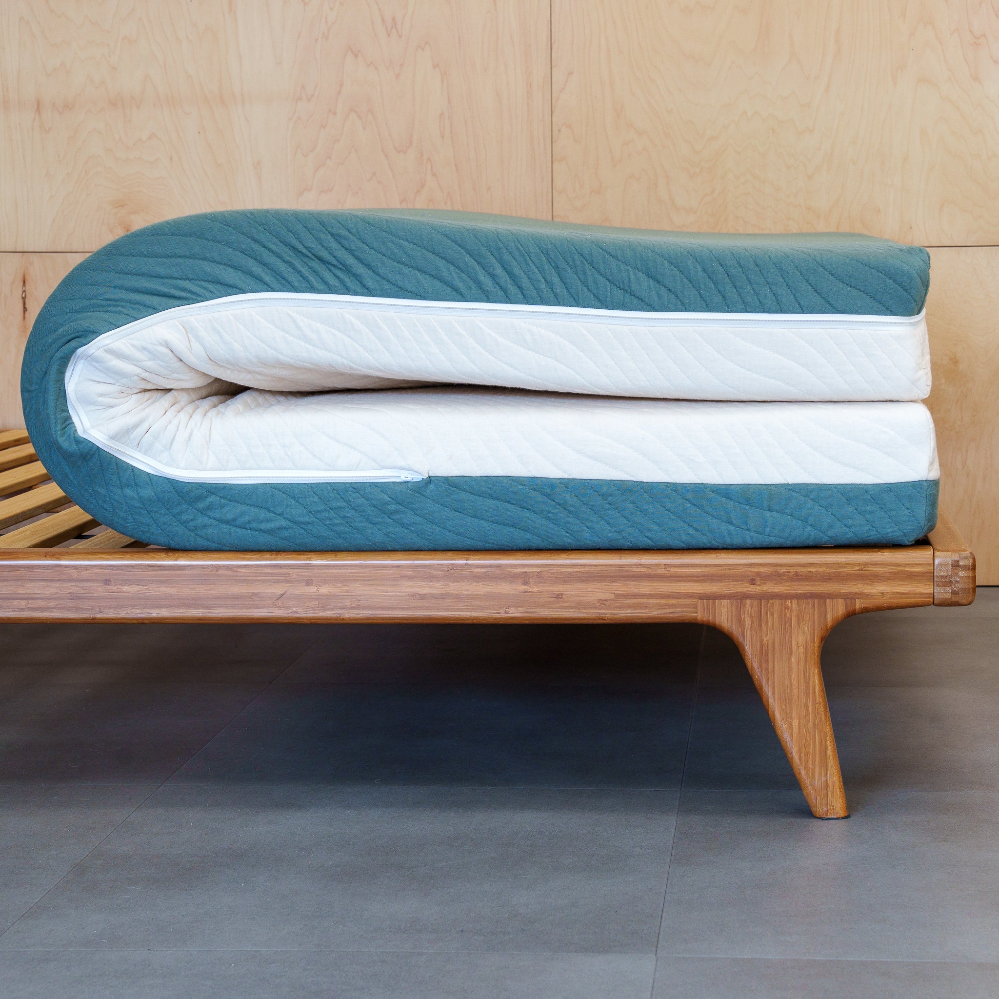 A folded tofino mattress with a quilted teal-blue exterior and white inner layer, resting on a wooden slatted bed frame against a light wood paneled wall.